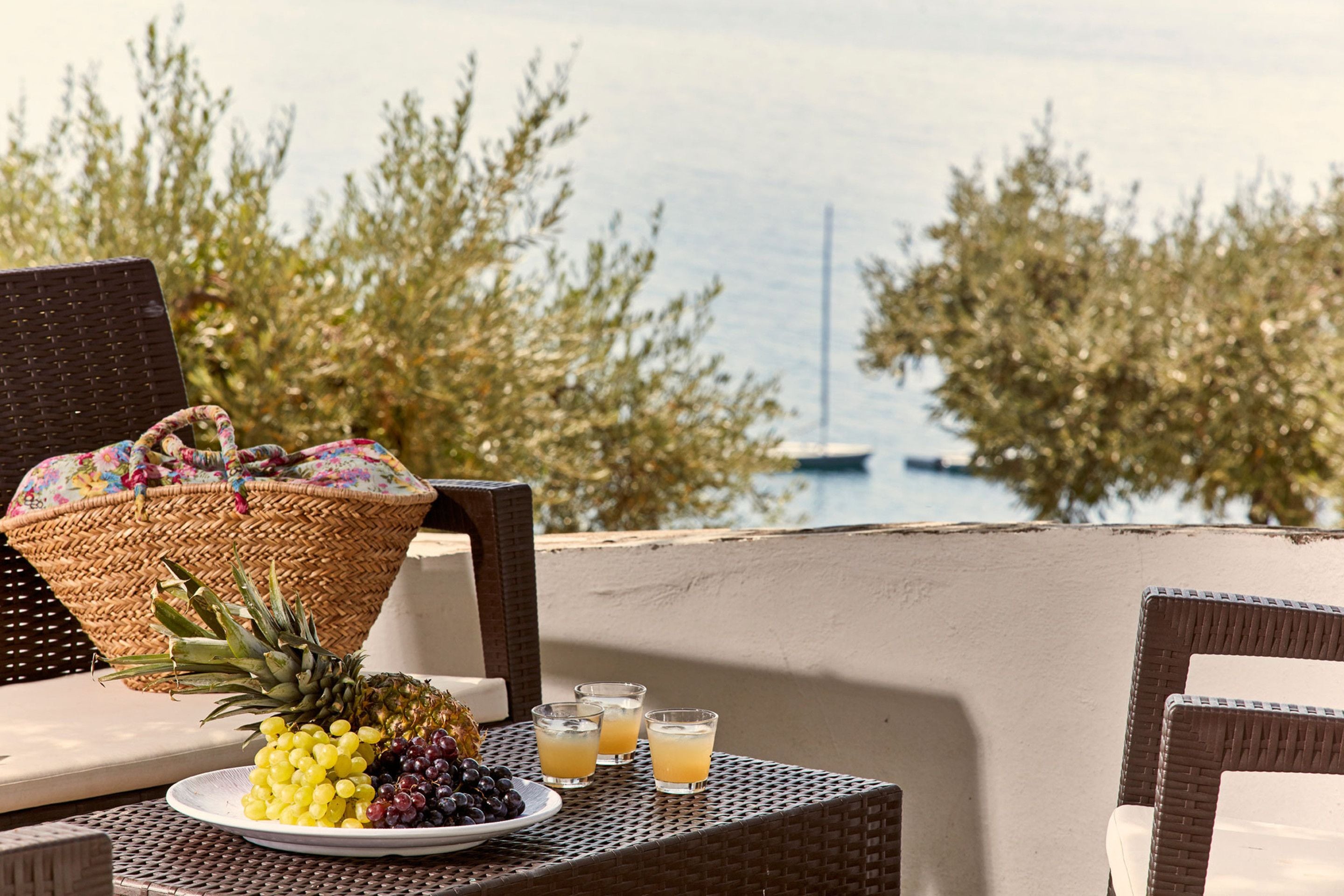 A wicker table with a plate of fruit and drinks, a wicker basket, and a view of the sea.