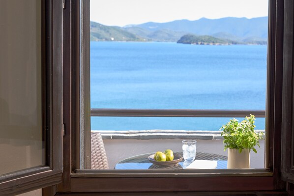 Vista desde una ventana sobre un cuerpo de agua con montañas a lo lejos, una mesa con un cuenco de fruta y un vaso de agua, y una planta en maceta.