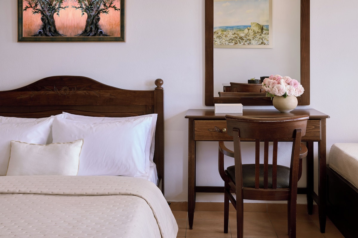 A neatly made bed with white linens, a wooden desk with a vase of flowers, and a chair.