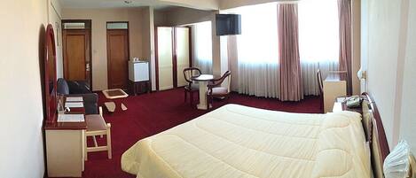 A hotel room with a large bed, a desk with a chair, a small table, a TV, and a window with curtains.