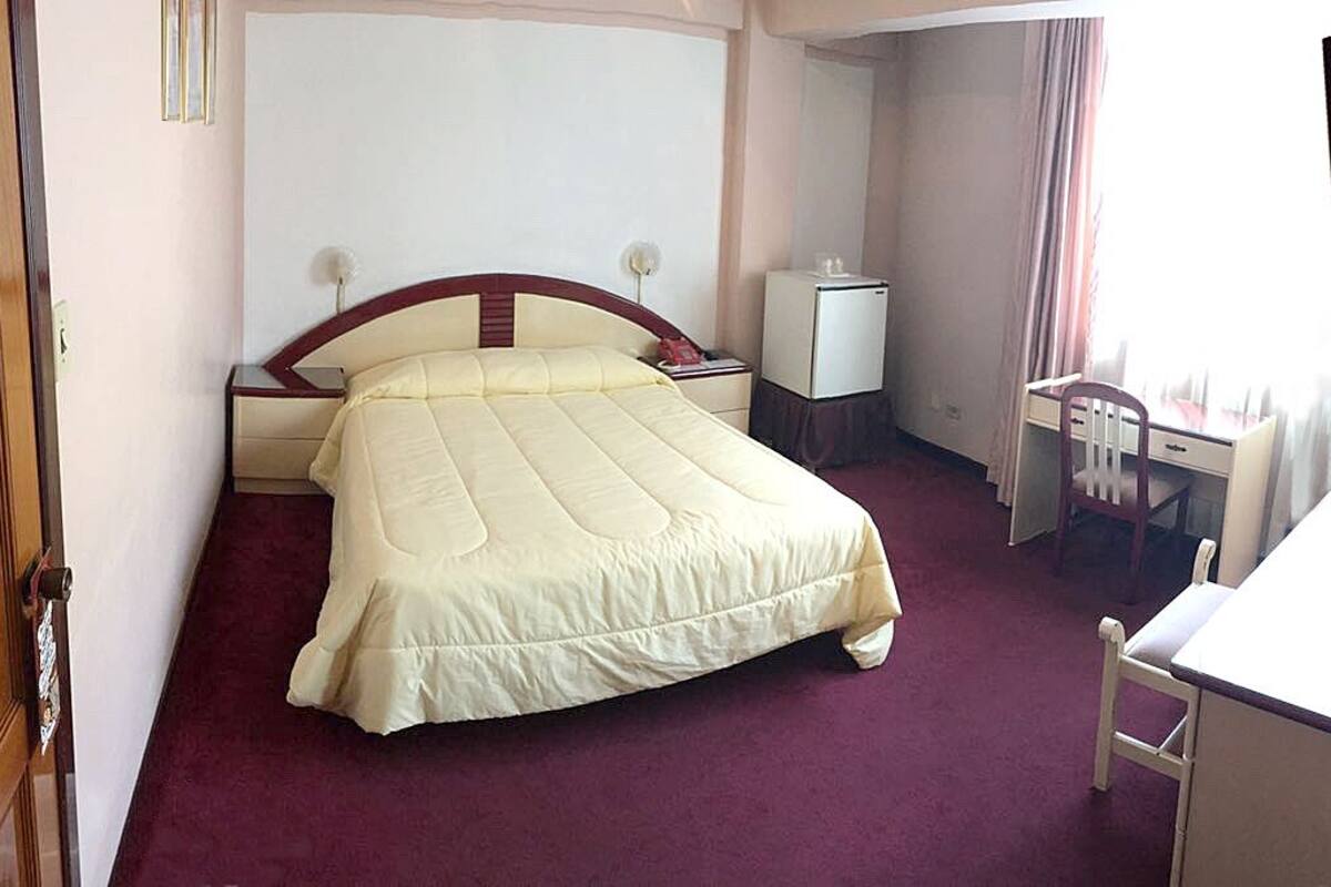A hotel room with a bed, bedside table, chair, desk, and a window with curtains.