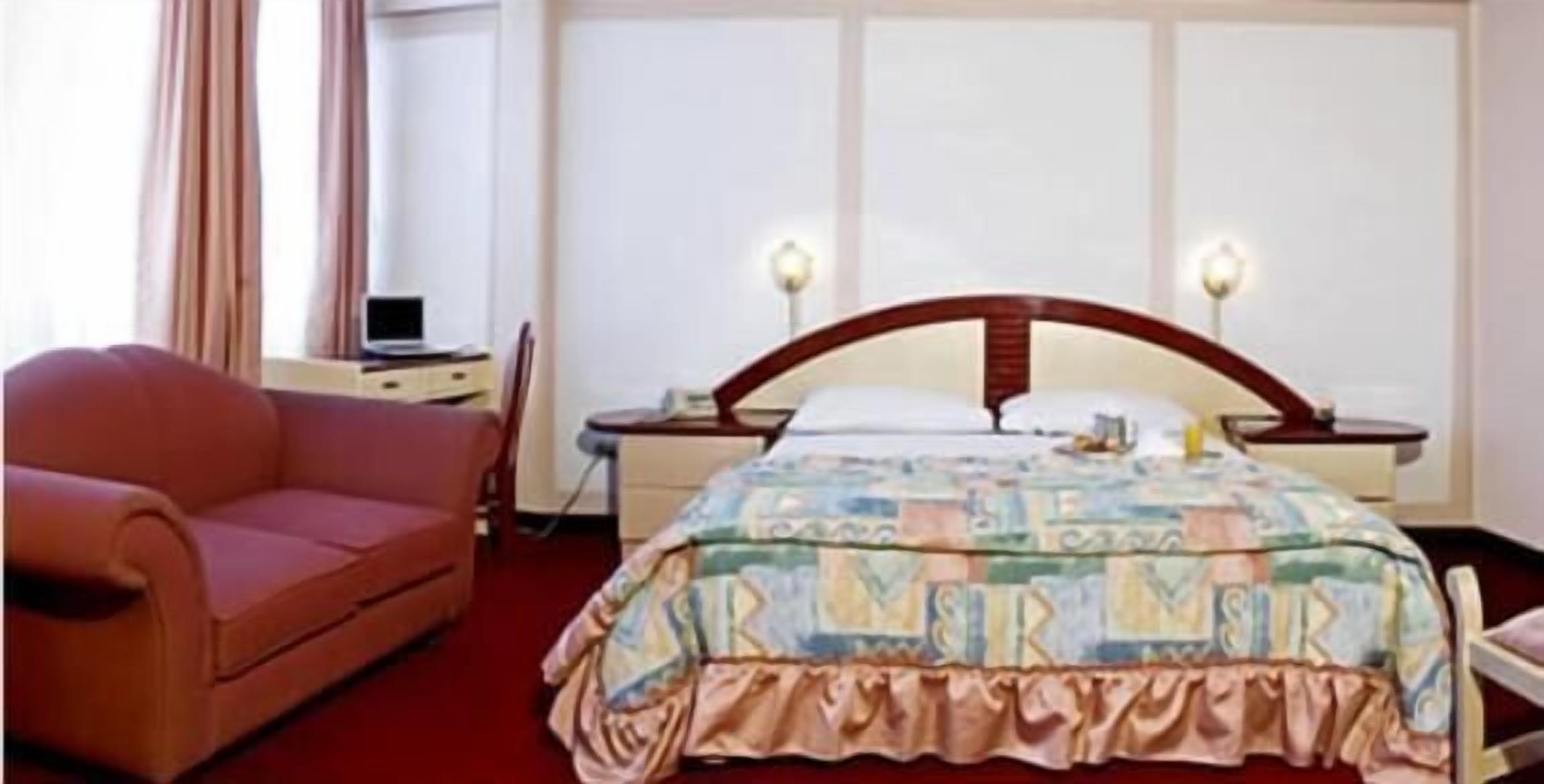 Een hotelkamer met een bed, een rode fauteuil, een nachtkastje met lamp en een raam met gordijnen.