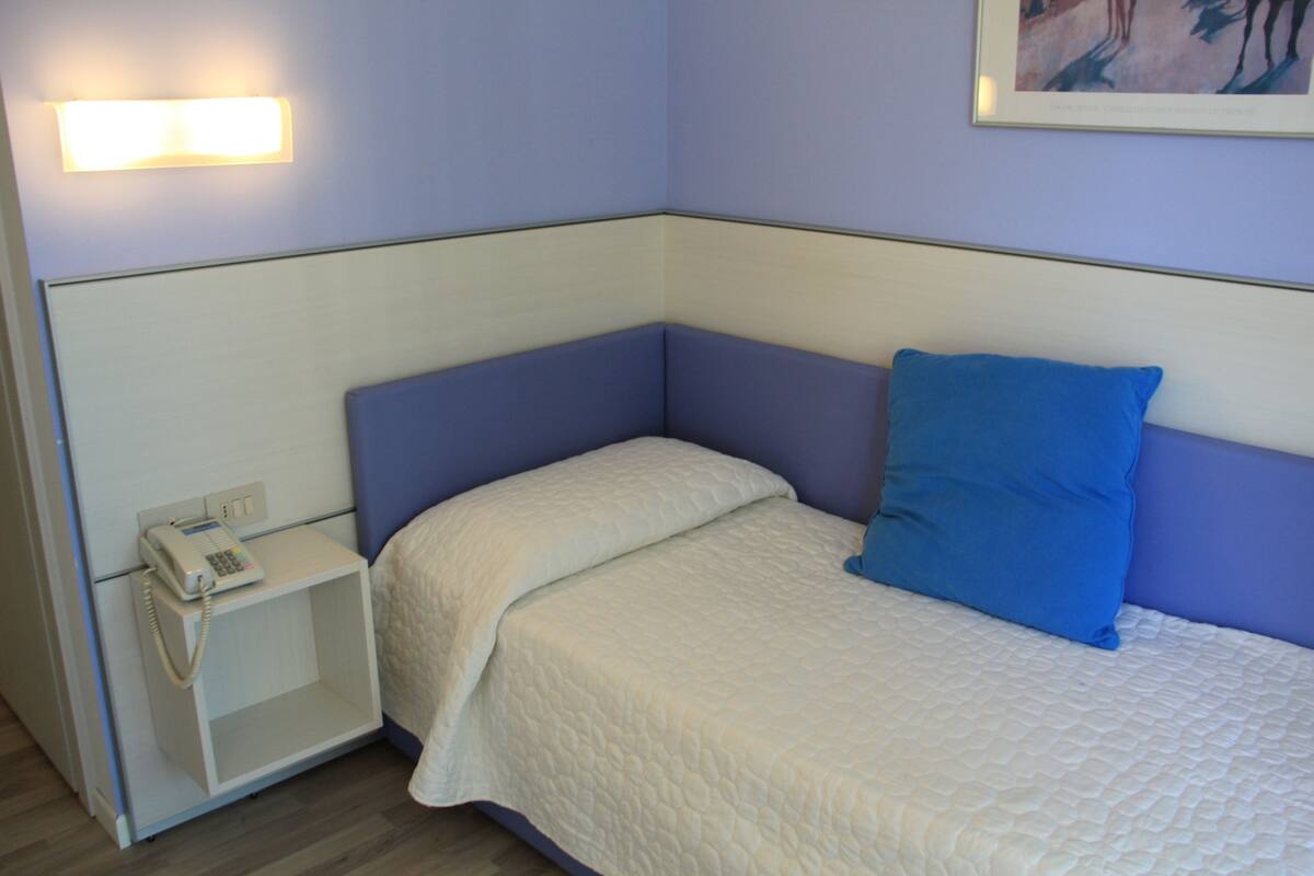 Une chambre d’angle avec un lit blanc, un oreiller bleu, une table de chevet avec un téléphone et une lampe fixée au mur.