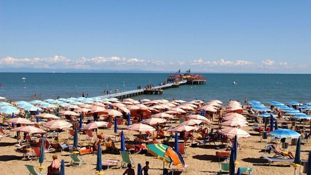A beach with numerous umbrellas and sun loungers, a pier extending into the sea, and a clear sky.