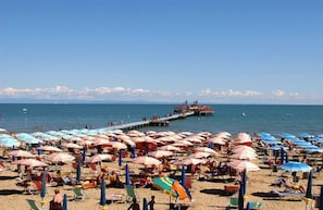 A beach with numerous umbrellas and sun loungers, a pier extending into the sea, and a clear sky.