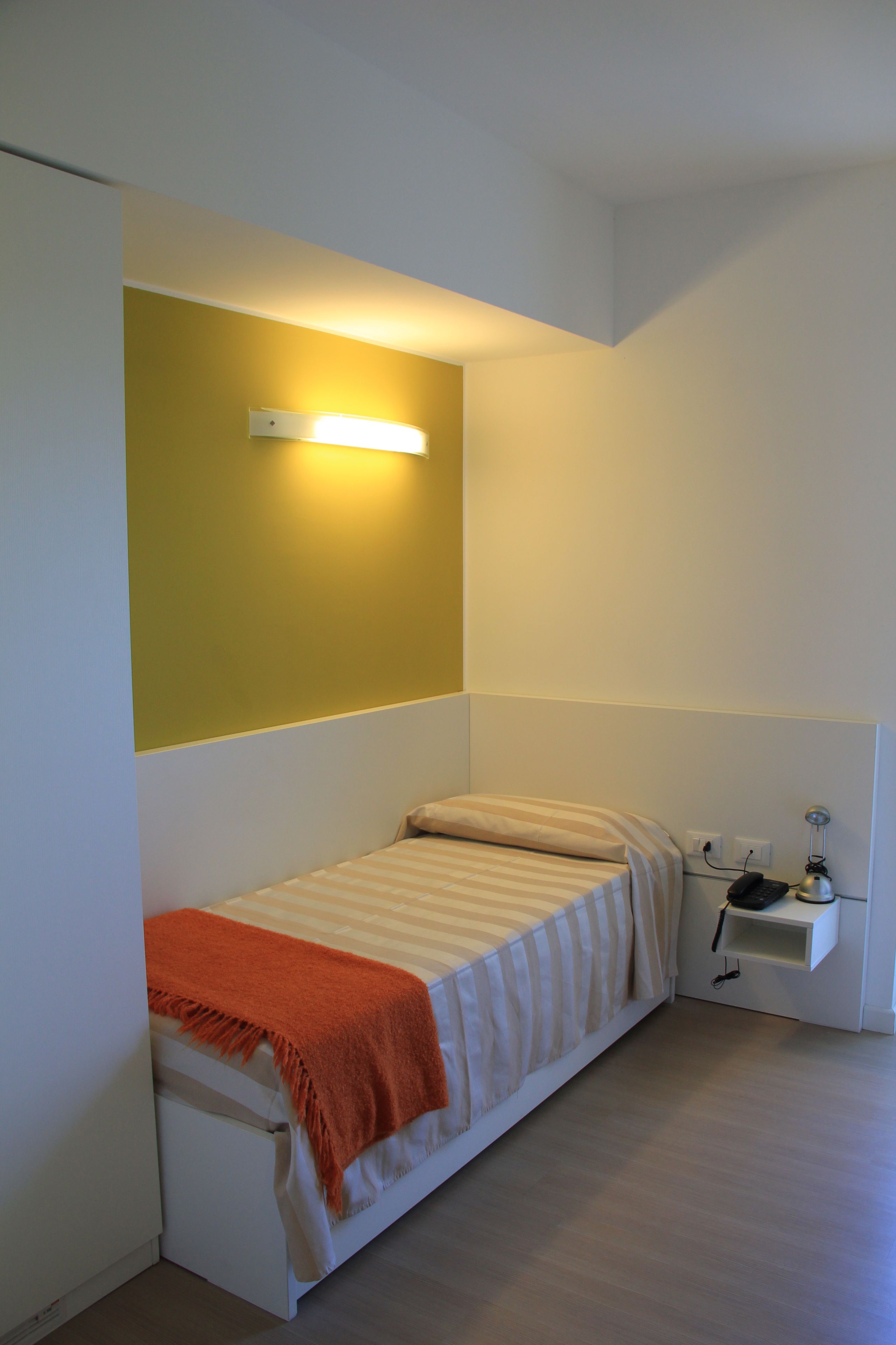 Quarto pequeno com uma cama de solteiro encostada a uma parede amarela, mesa de cabeceira com abajur e uma tomada.