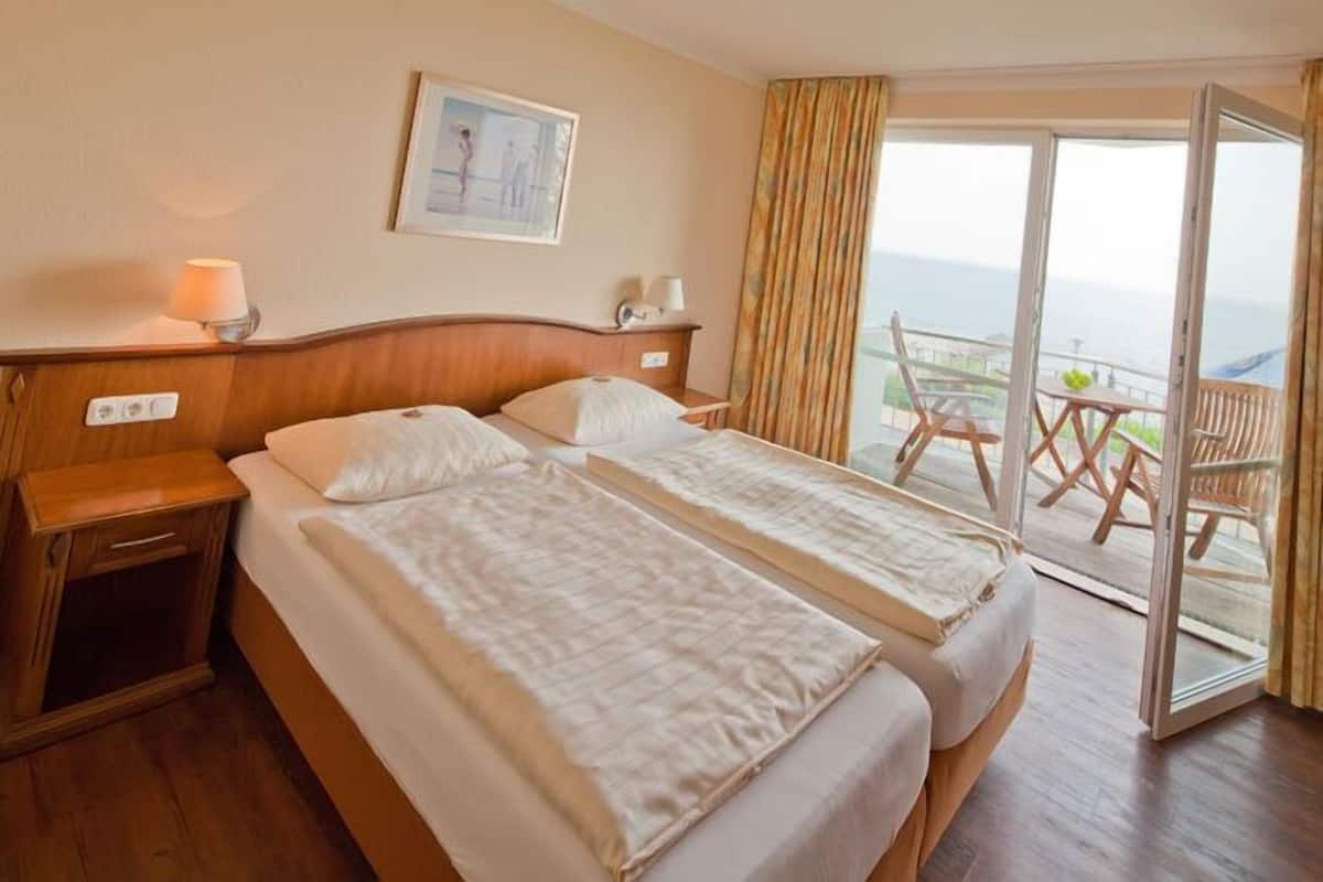 Une chambre d’hôtel avec deux lits, une tête de lit en bois, une table de chevet, une lampe, un tableau encadré au mur et un balcon avec vue.