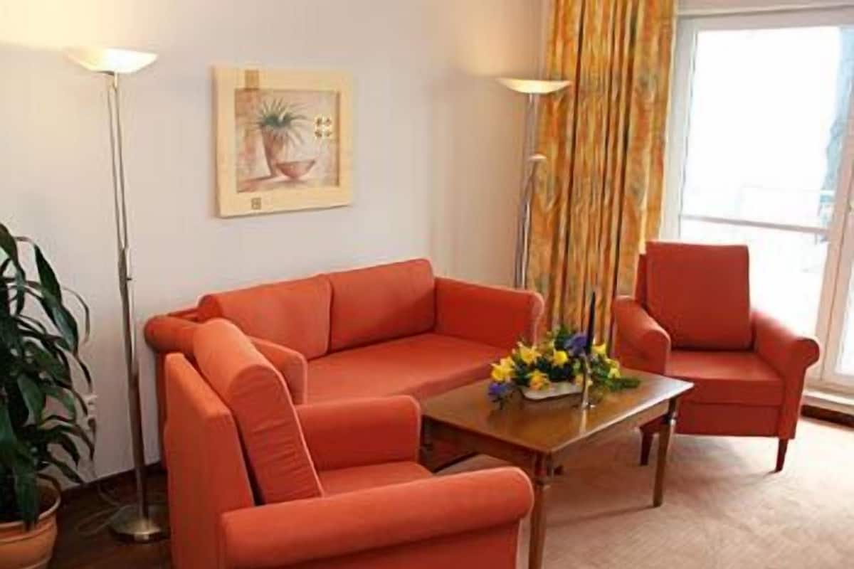 Un salon avec un canapé rouge, un fauteuil assorti, une table basse ornée de fleurs, un lampadaire et un tableau accroché au mur.