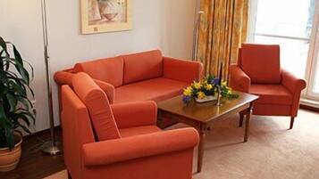 Uma sala de estar com um sofá vermelho, uma poltrona combinando, uma mesa de centro com flores, um abajur e um quadro na parede.