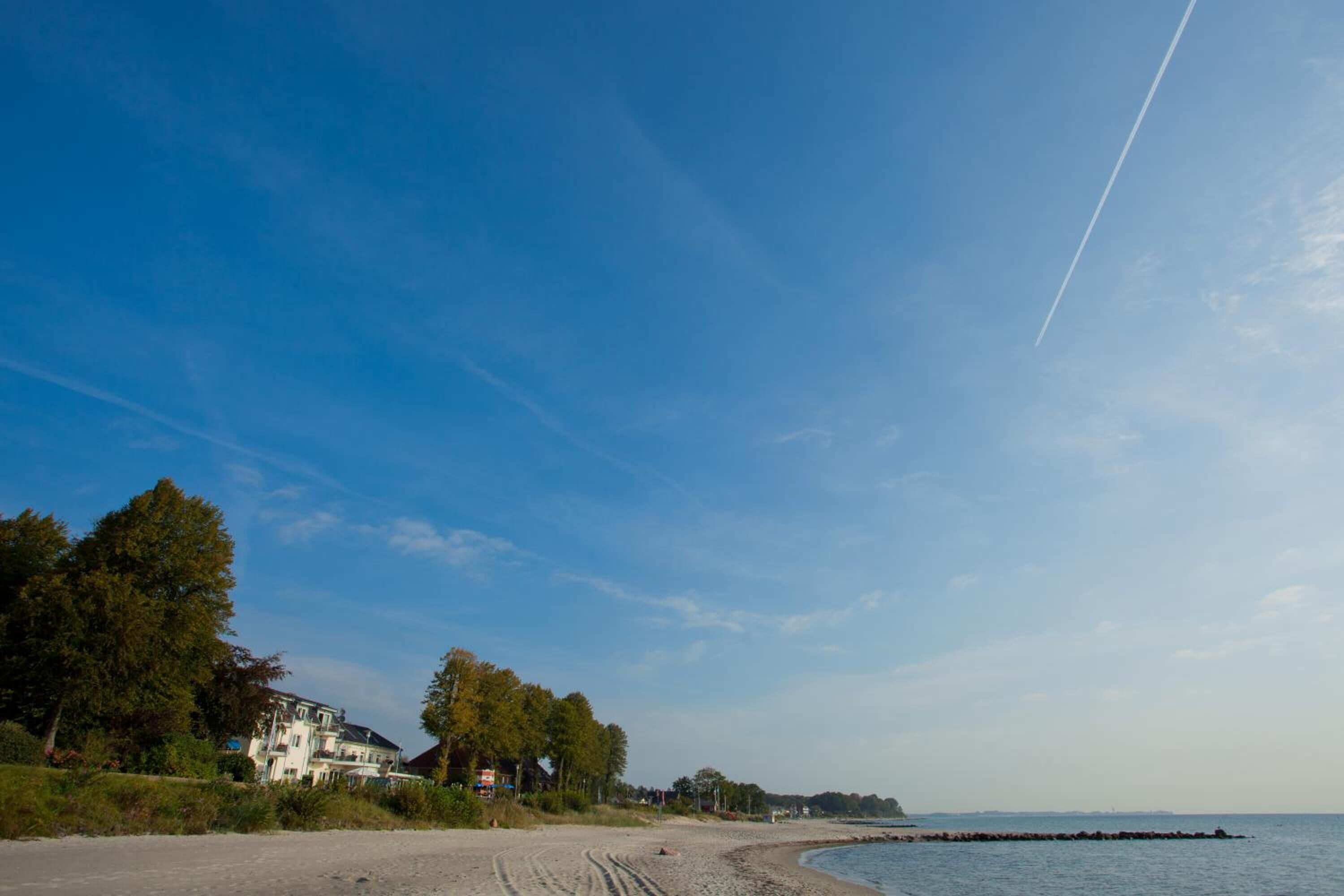 Ein Strand mit einem großen Haus links, blauem Himmel und einem sichtbaren Kondensstreifen.