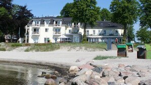 Hotel de varios pisos con balcones, situado cerca de una masa de agua con playa de arena y rocas.