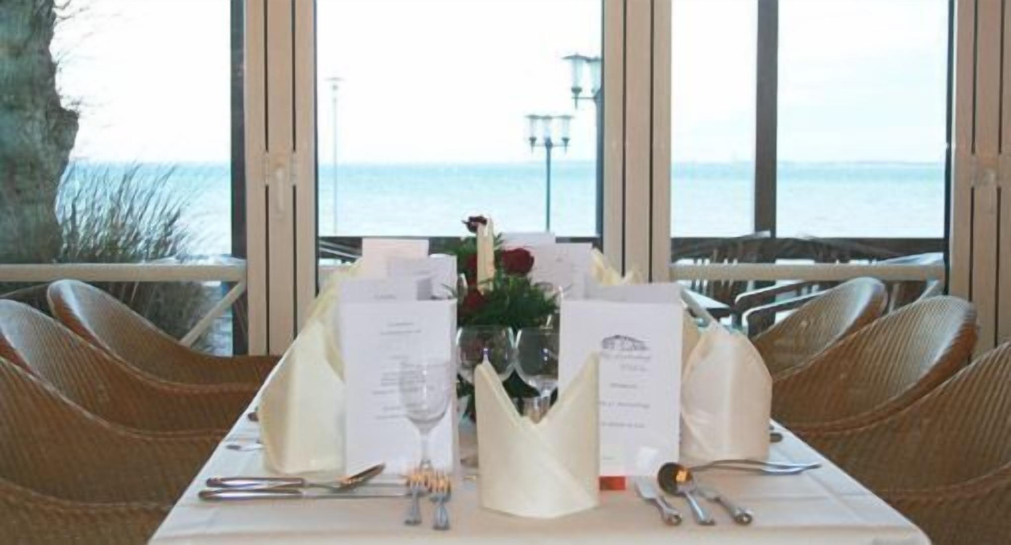 A dining table set for two with menus, a vase with flowers, and a view of the sea through large windows.