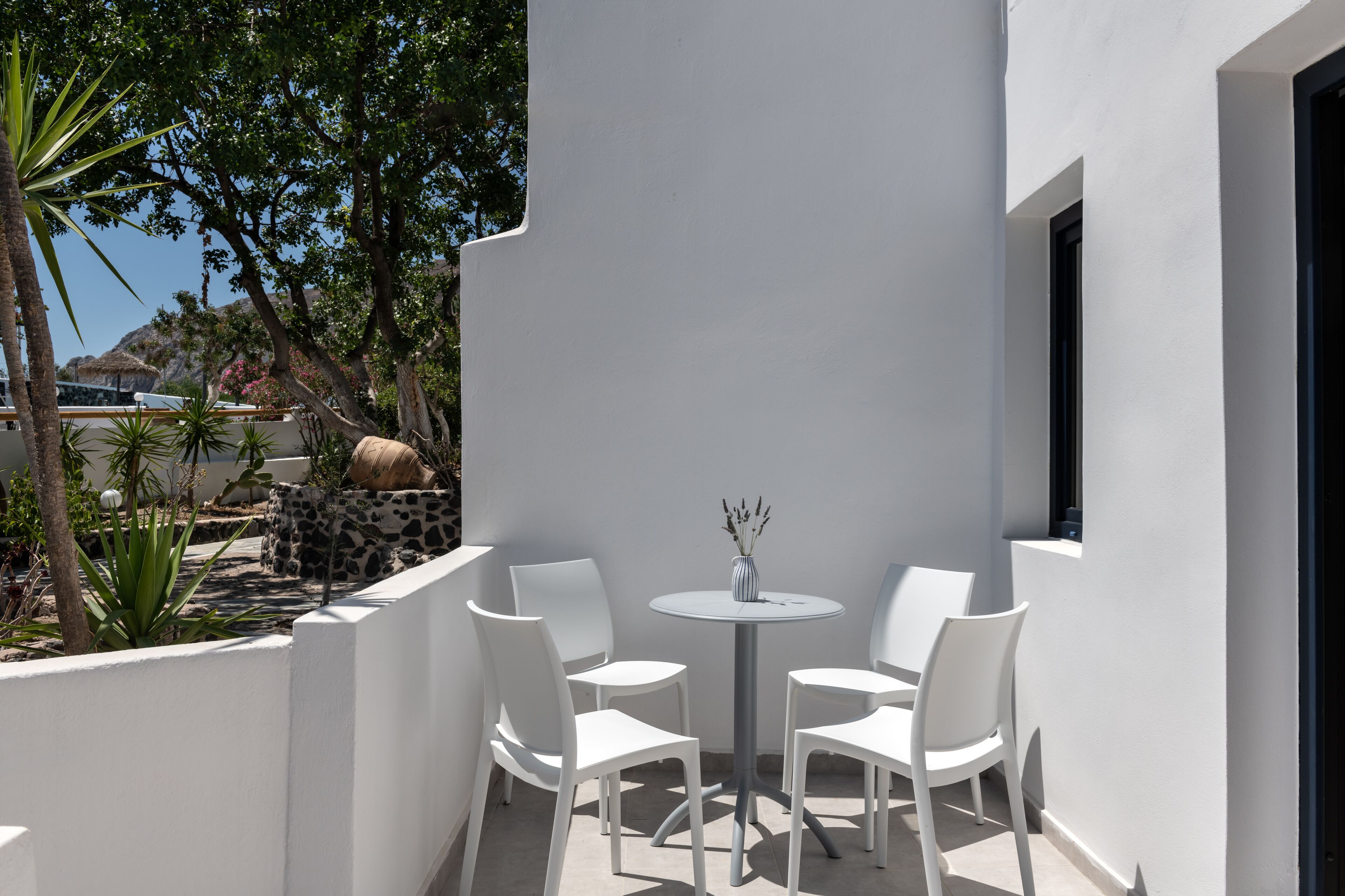 Un espace extérieur aménagé avec des sièges blancs, comprenant une table ronde et quatre chaises, entouré d’un mur blanc et d’un petit jardin agrémenté de plantes et d’un rocher.