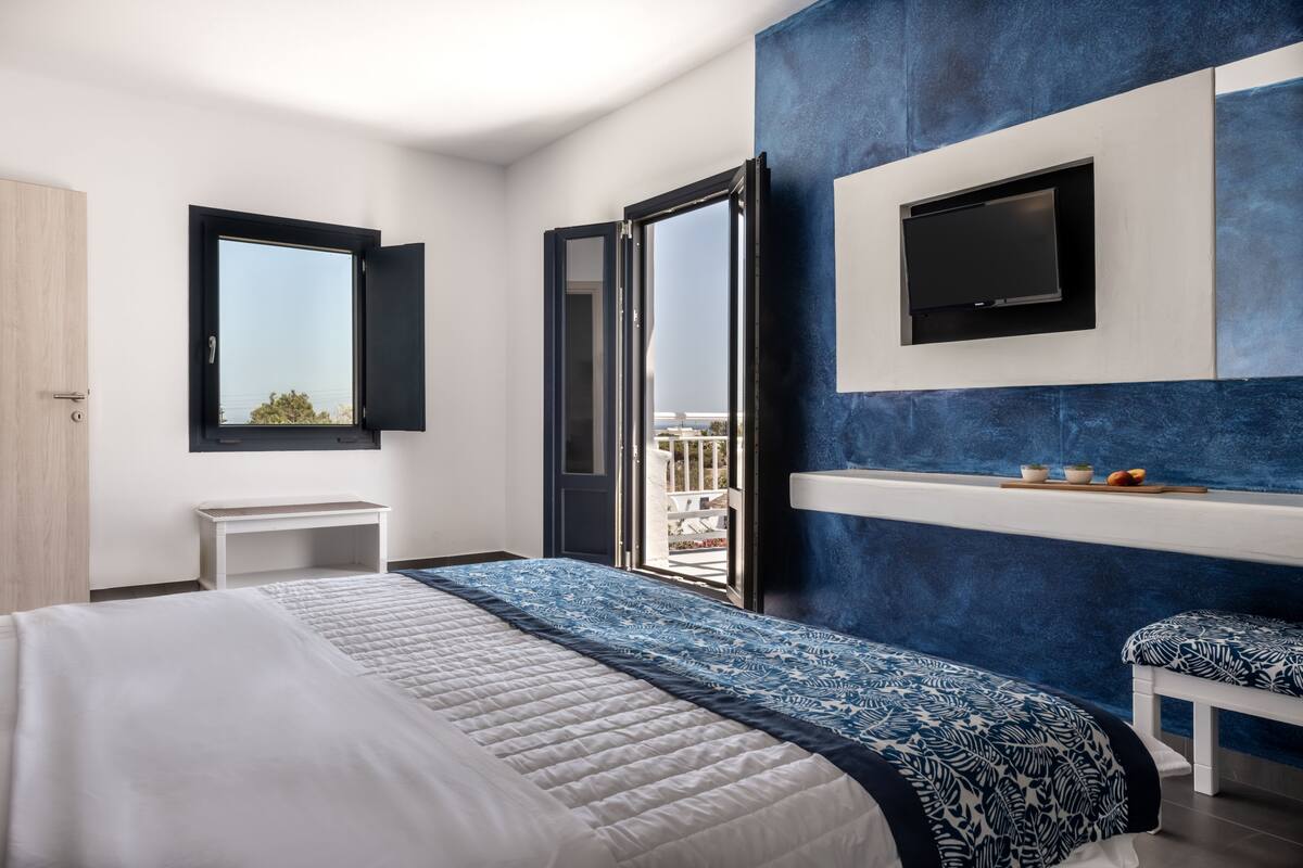 Un dormitorio moderno con una cama grande, un televisor instalado en la pared y una ventana con puerta abierta que da al exterior.