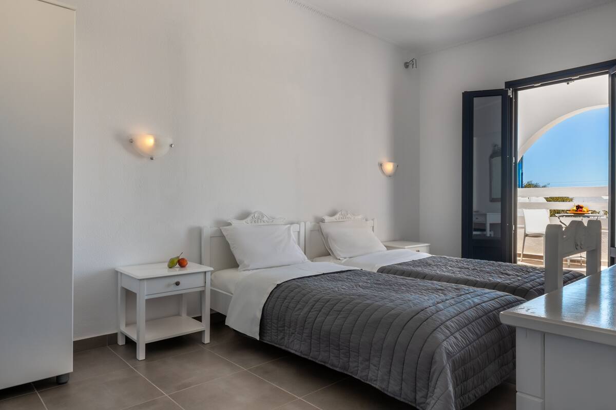Un dormitorio con dos camas, una mesita de noche con fruta y vista al mar por una puerta abierta.