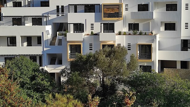 A modern multi-story building with balconies, large windows, and a stone wall in the background. - Amorani Studios (Andros)