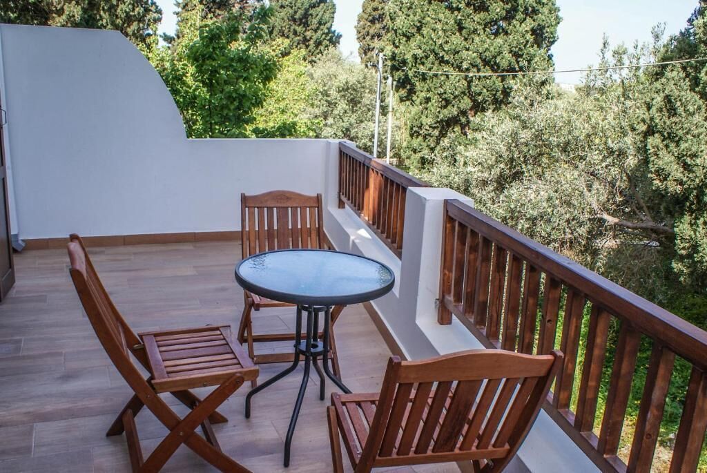 A balcony with wooden furniture and a glass table, surrounded by a white wall and a wooden railing, overlooking a green landscape.