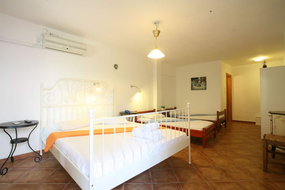 A room with a white bed, a small table, and a chair. There is a wall-mounted air conditioner and a painting on the wall.