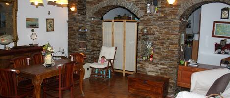 Un coin repas avec une table et des chaises en bois, un fauteuil blanc et un mur en briques agrémenté d’une porte en arc de cercle.