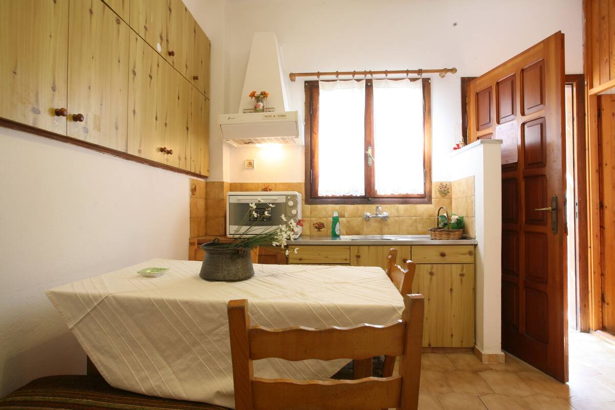 A kitchen with wooden cabinets, a dining table with a tablecloth, a window with curtains, and a sink.