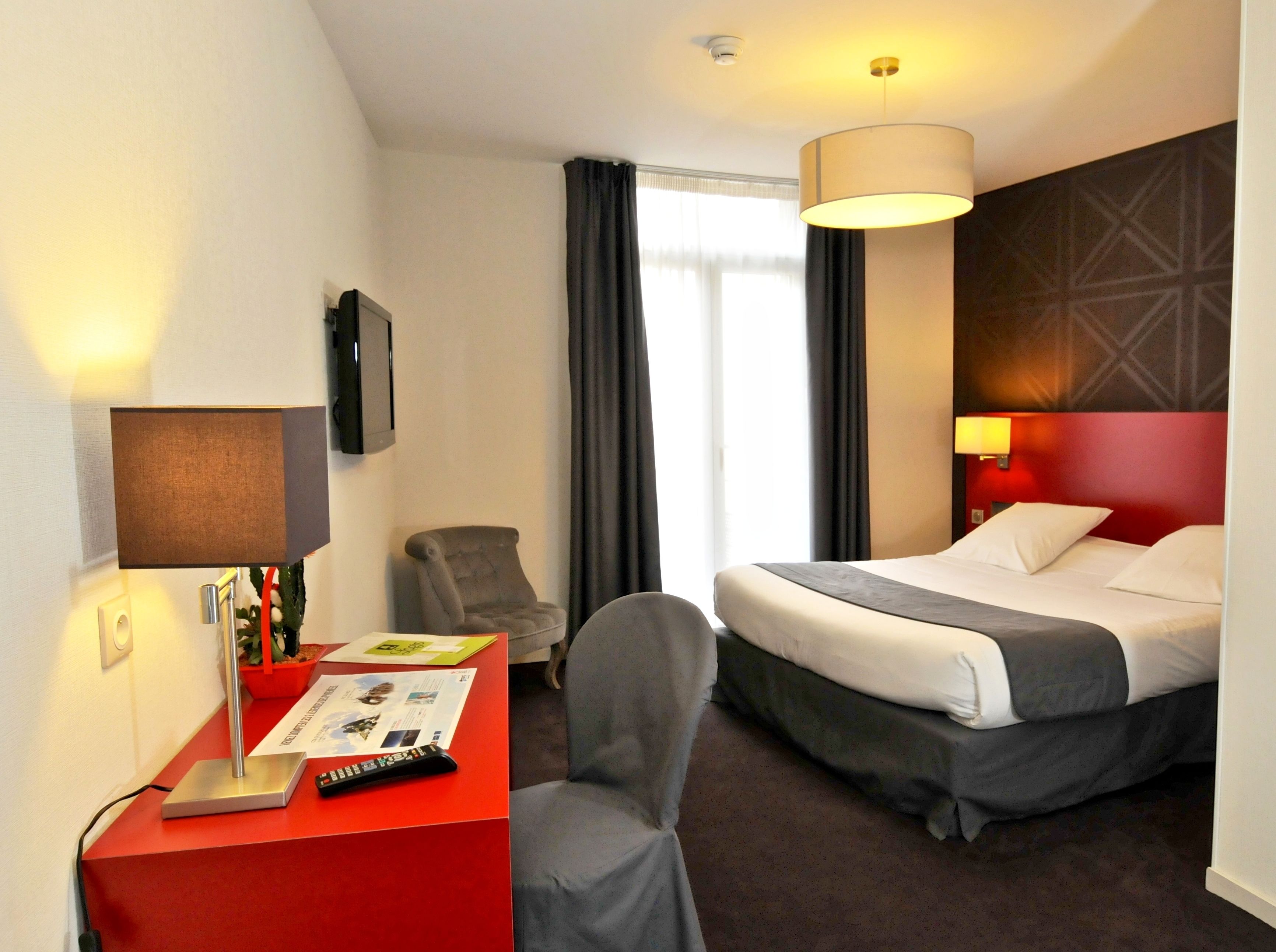 A hotel room with a bed, a desk with a red chair, a TV, and a window with curtains.