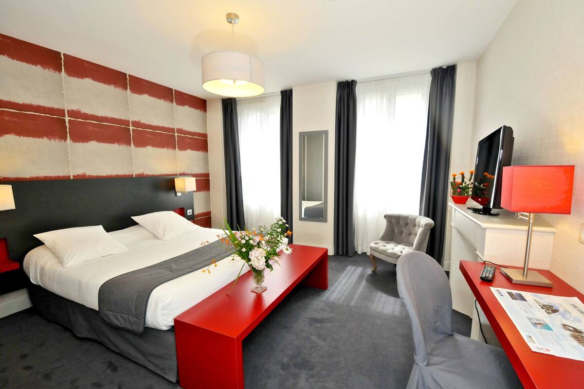 A hotel room with a large bed, a desk with a red lamp, a chair, a TV, and a vase with flowers.