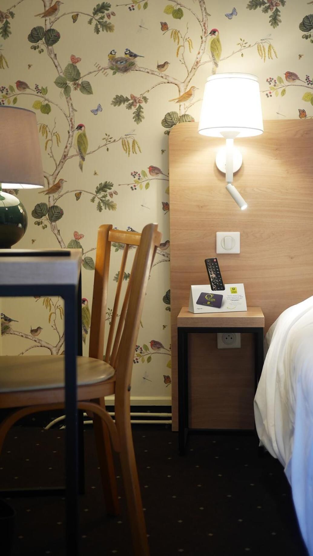 A hotel room with a wooden headboard, a bedside table with a lamp, a remote control, and a card on the table.