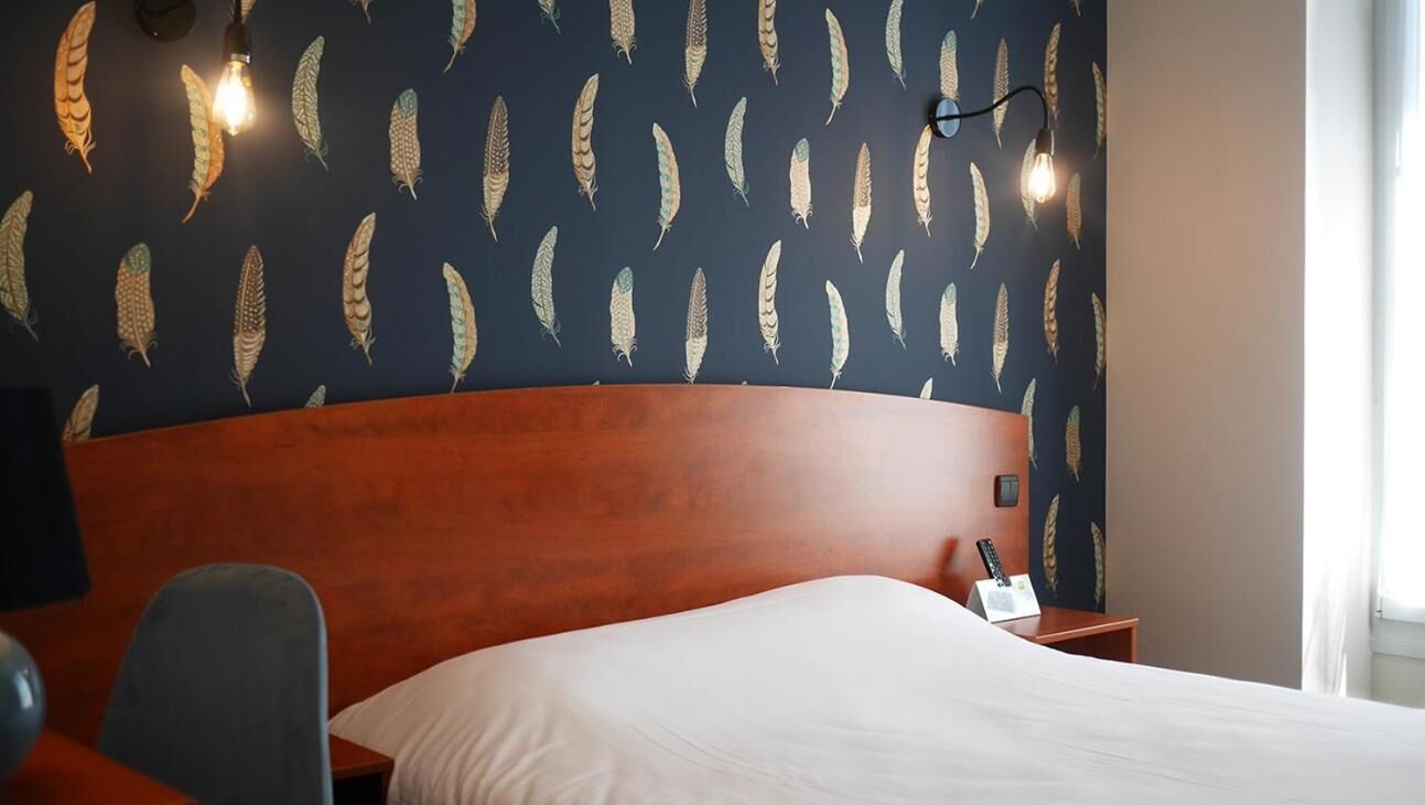 Ein Hotelzimmer mit dunkel gemustertem Wandteppich, einem Holz-Kopfteil, einem weißen Bett, einem Schreibtisch mit Lampe und einem Sessel.