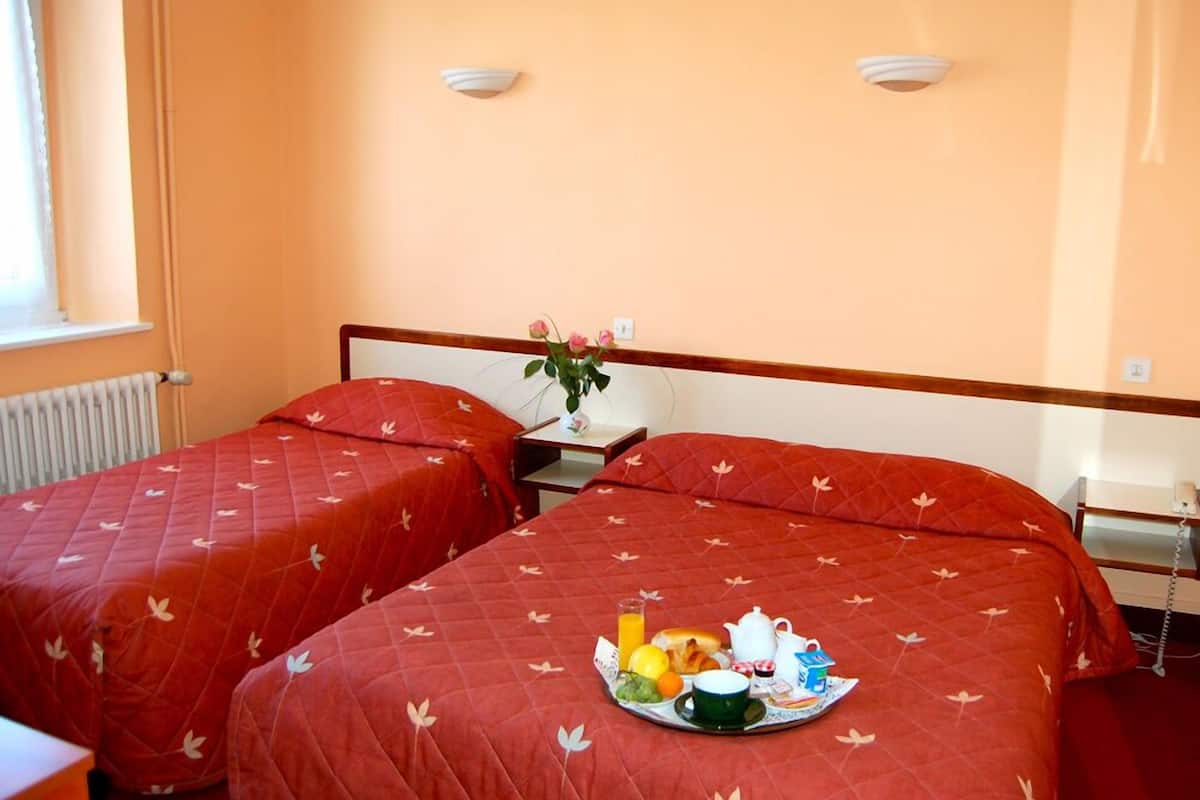 Ein Hotelzimmer mit zwei Betten, beide mit roter Bettdecke mit Blättermotiv. Auf dem mittleren Bett steht ein Tablett mit Frühstücksgebäck.