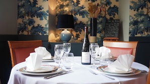 Una mesa de comedor dispuesta con platos, vasos y una botella de vino, rodeada de sillas y una lámpara decorativa.