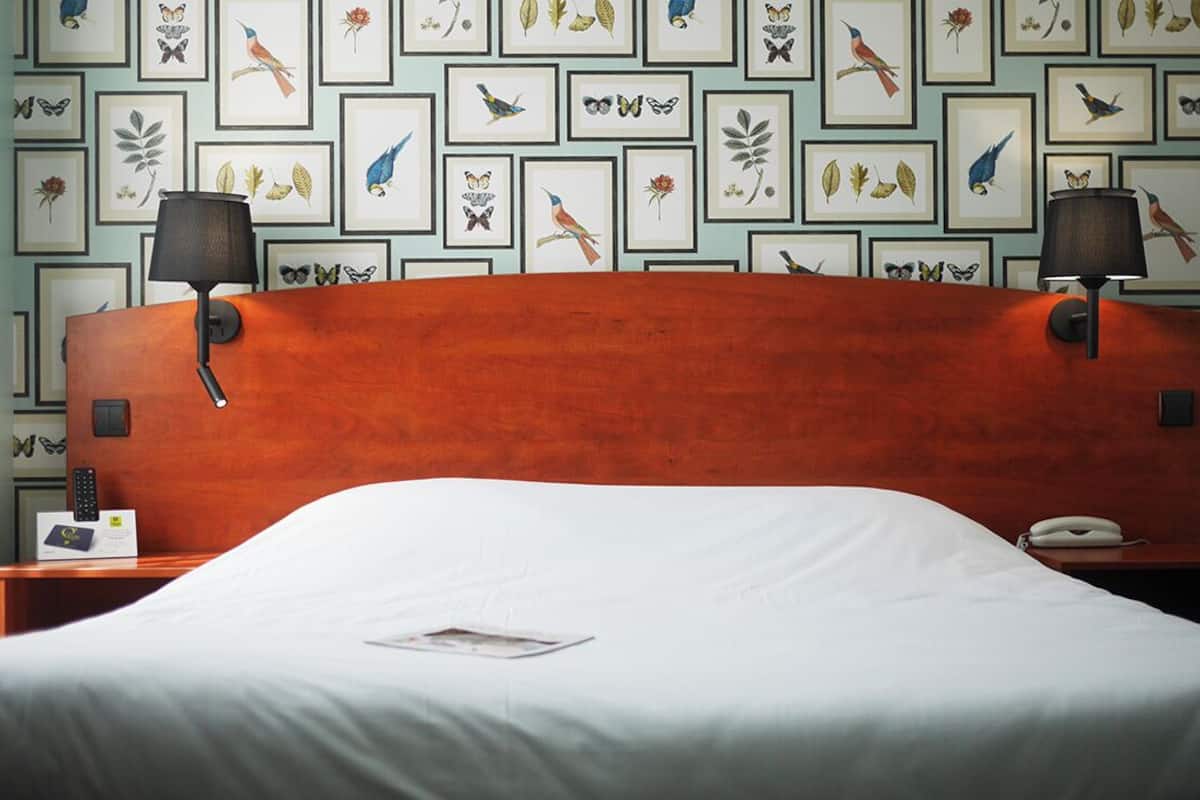 Ein Hotelzimmer mit einem hölzernen Kopfteil, zwei Nachttischlampen, einer weißen Bettdecke und gerahmten Pflanzenbildern an der Wand.