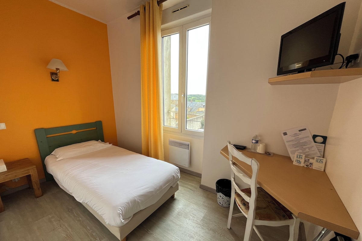 Ein Hotelzimmer mit einem Bett, einem Schreibtisch mit Stuhl, einem Fernseher und einem Fenster mit Vorhängen.