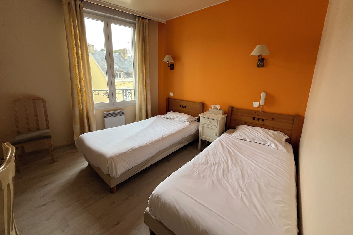 Ein Hotelzimmer mit zwei Einzelbetten, einem Sessel, einem Nachttisch mit Lampe und Blick auf Gebäude durch ein Fenster.
