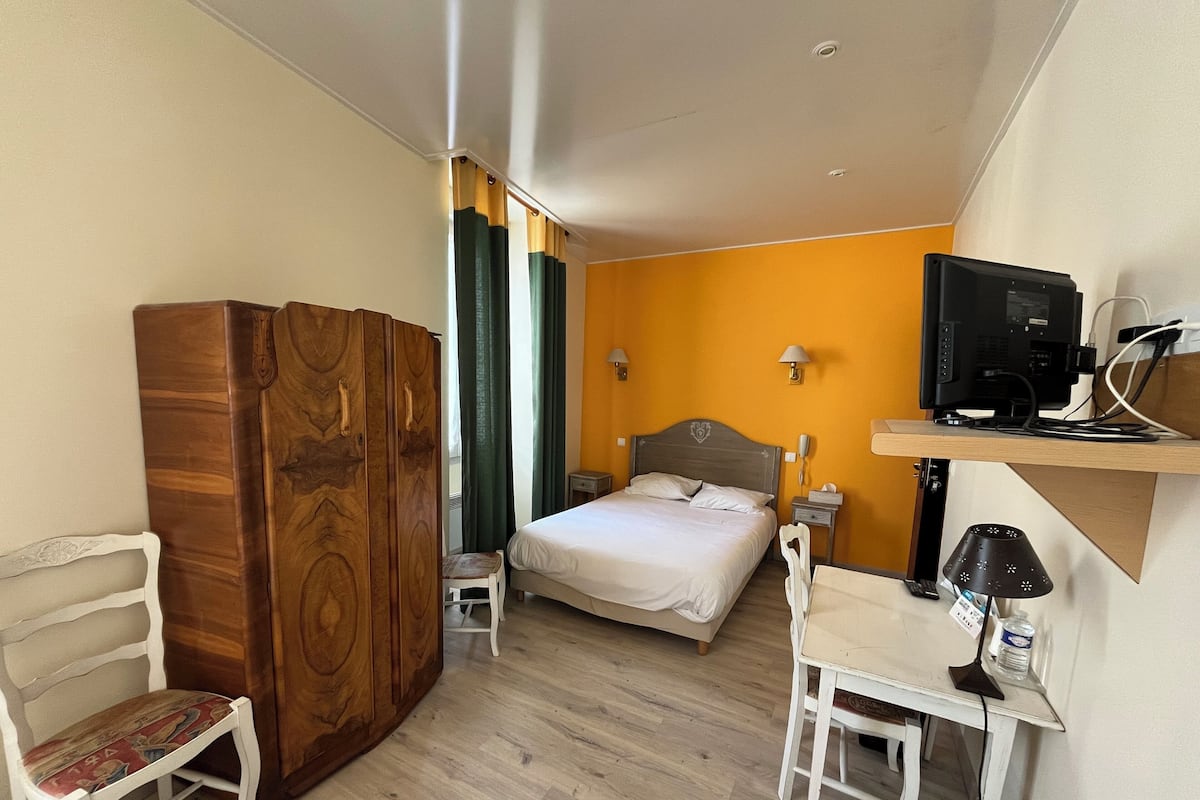 Ein Hotelzimmer mit Bett, Schreibtisch, Stuhl, Kleiderschrank, Fernseher und einem kleinen Tisch mit Lampe.