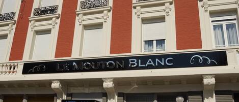 A hotel facade with a sign reading "LE MOUTON BLANC" and a "HOTEL" sign below it.