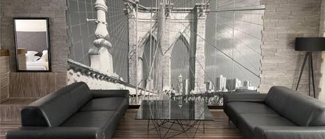 A modern living room with a large black and white photograph of a bridge, two black sofas, a glass coffee table, and a black floor lamp.