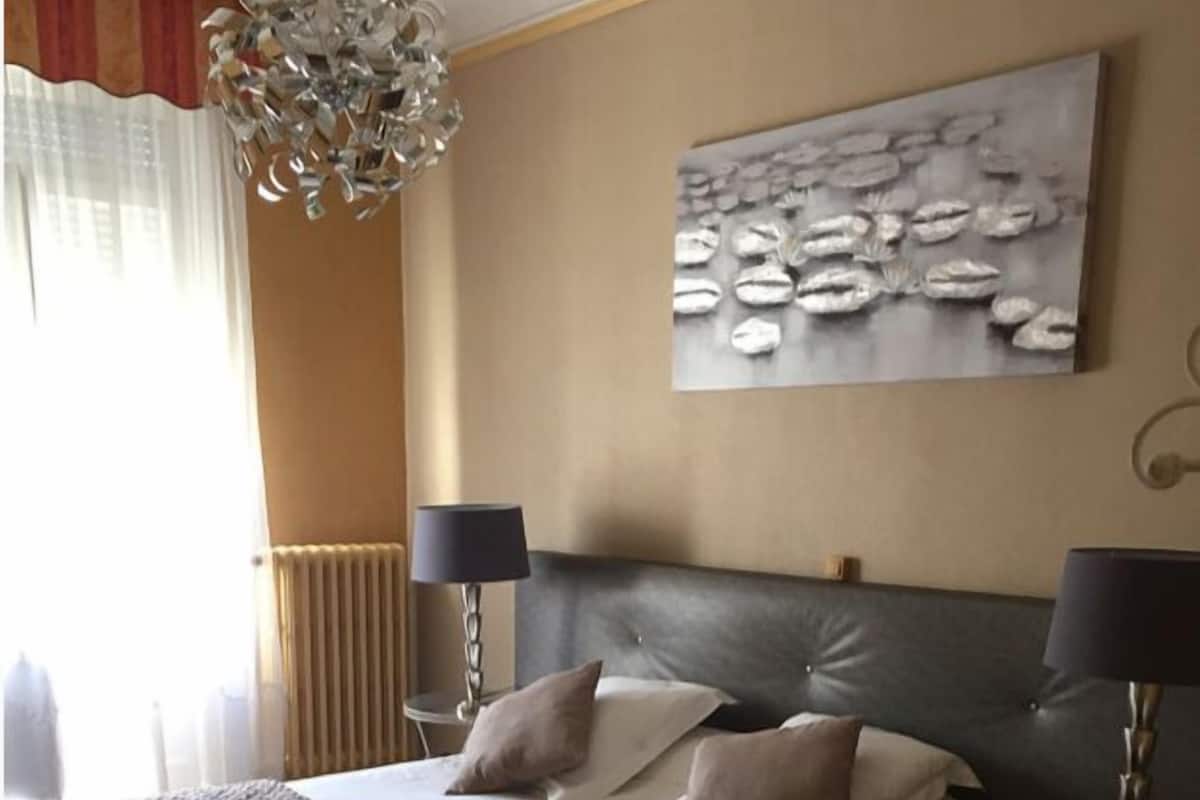A hotel room with a large bed, two bedside tables with lamps, a wall-mounted artwork, and a decorative chandelier.