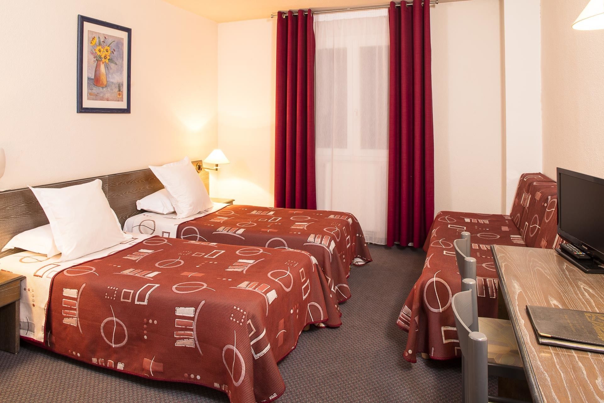 A hotel room with two beds, a desk, a television, and a window with red curtains.