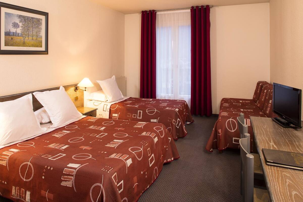 A hotel room with two beds, a desk with a television, and a window with red curtains.