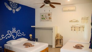 Quarto com duas camas, ventilador de teto, ar condicionado, decorações nas paredes e uma pequena mesa com abajur.