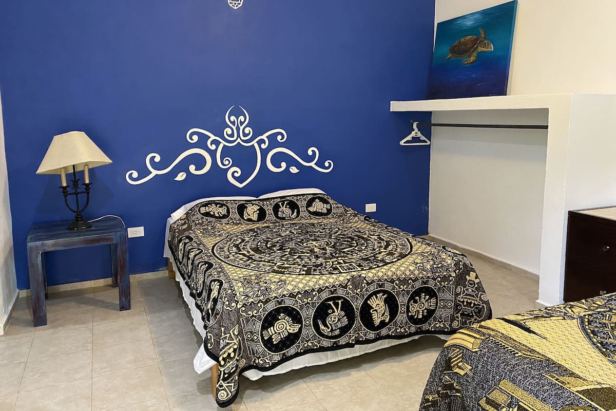 Un dormitorio con pared azul, cama con colcha estampada, mesita de noche con lámpara y un cuadro de una tortuga.