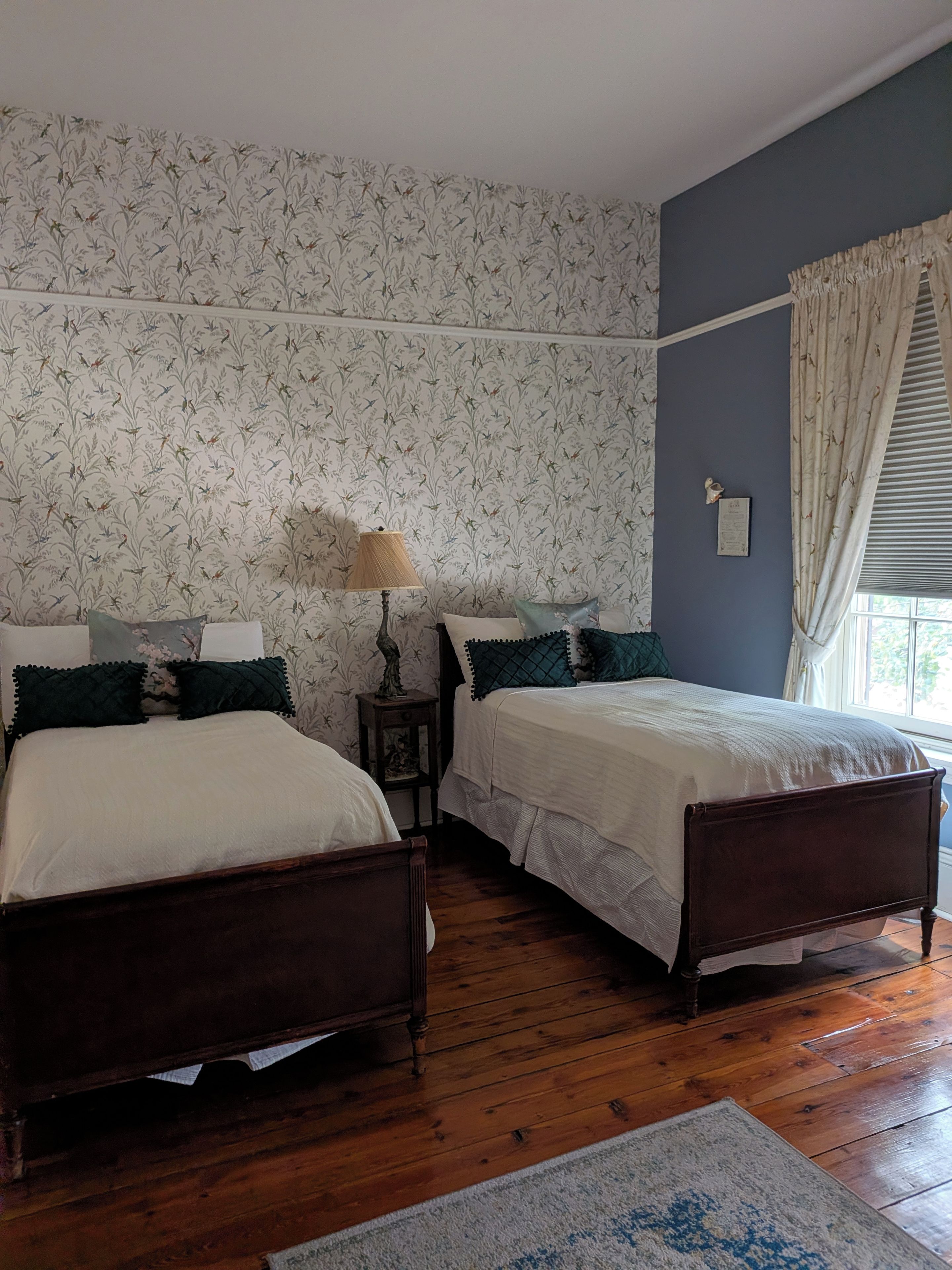 A bedroom with two wooden beds, a nightstand with a lamp, floral wallpaper, a window with curtains, and a rug on the wooden floor.