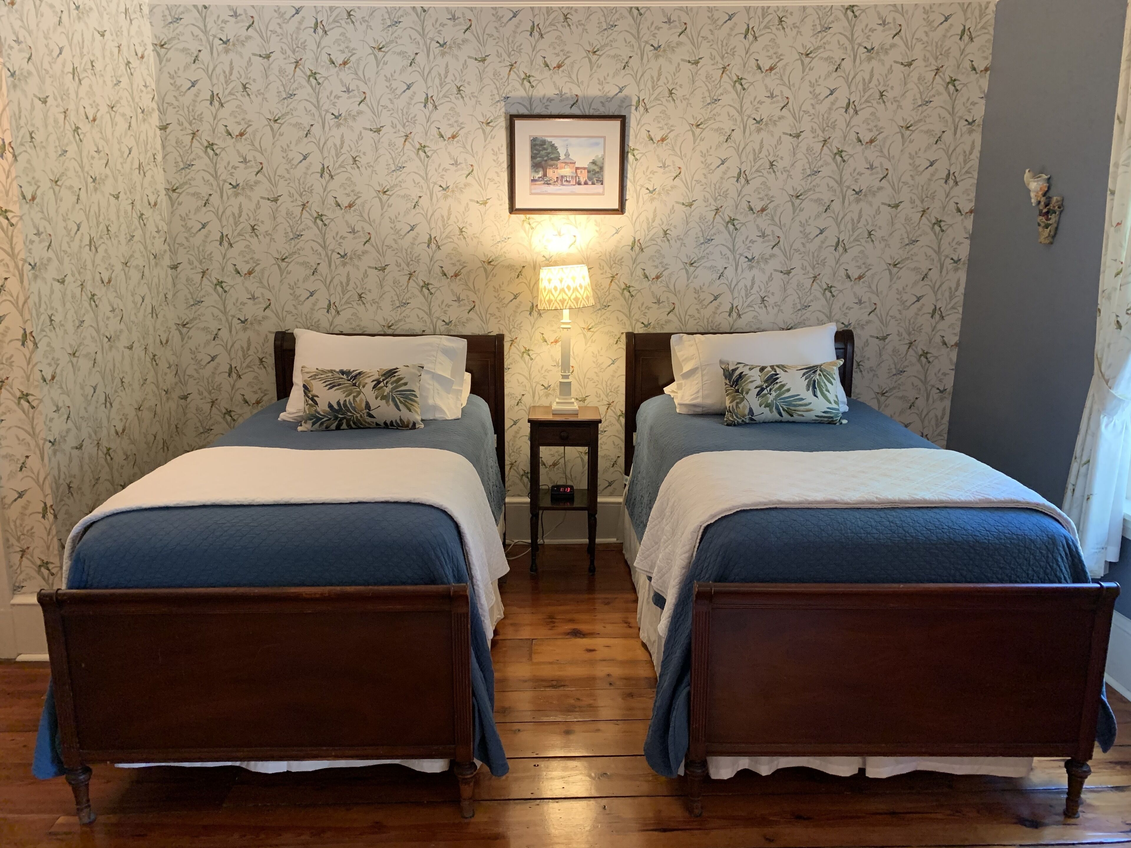 Quarto com duas camas de madeira, cada uma com colcha azul e travesseiro branco. Há uma pequena mesa com abajur entre as camas. As paredes têm um papel de parede com padrão floral.