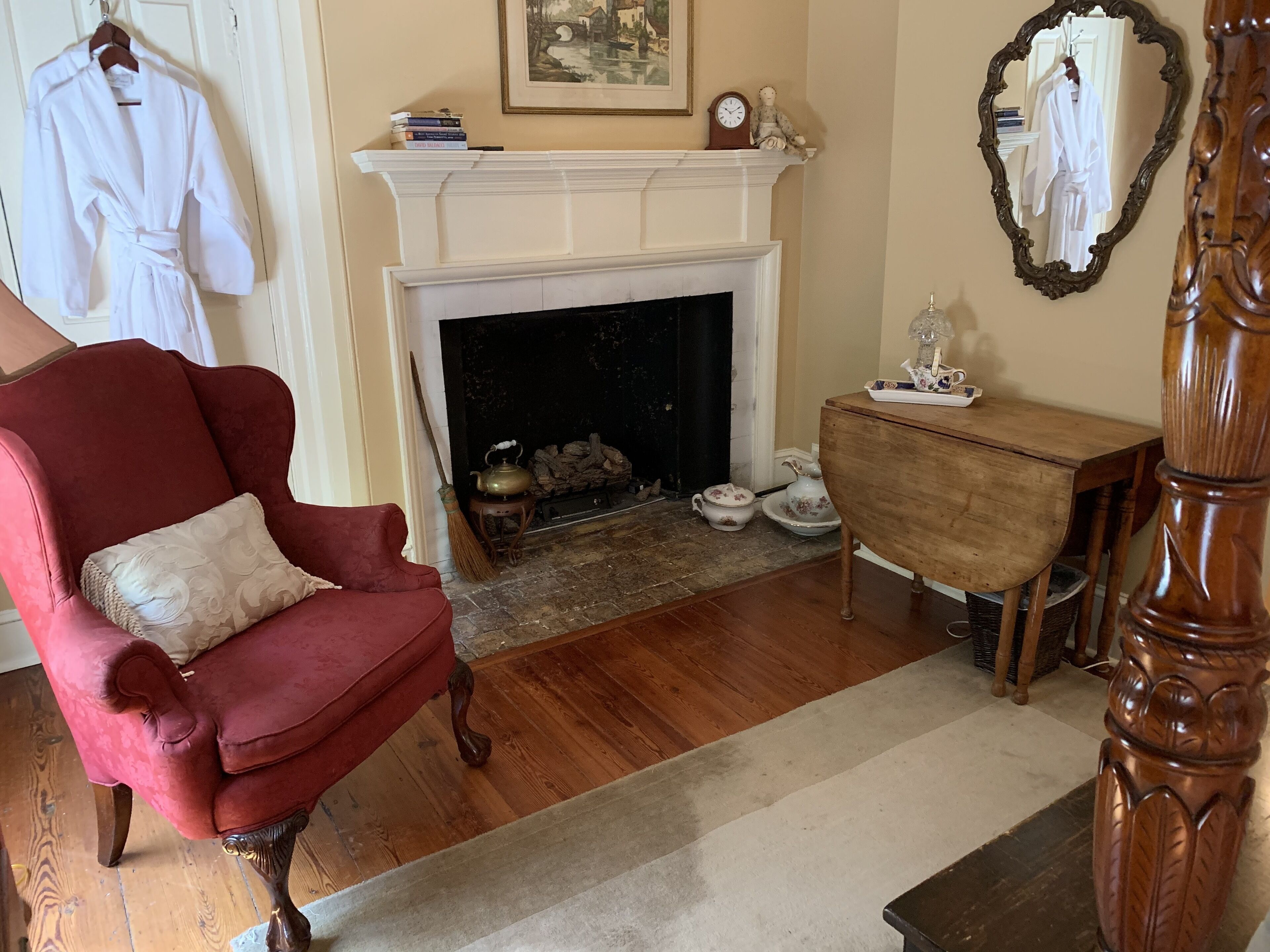 A room with a red armchair, a white fireplace, a wooden side table, and a mirror with a white robe hanging on it.