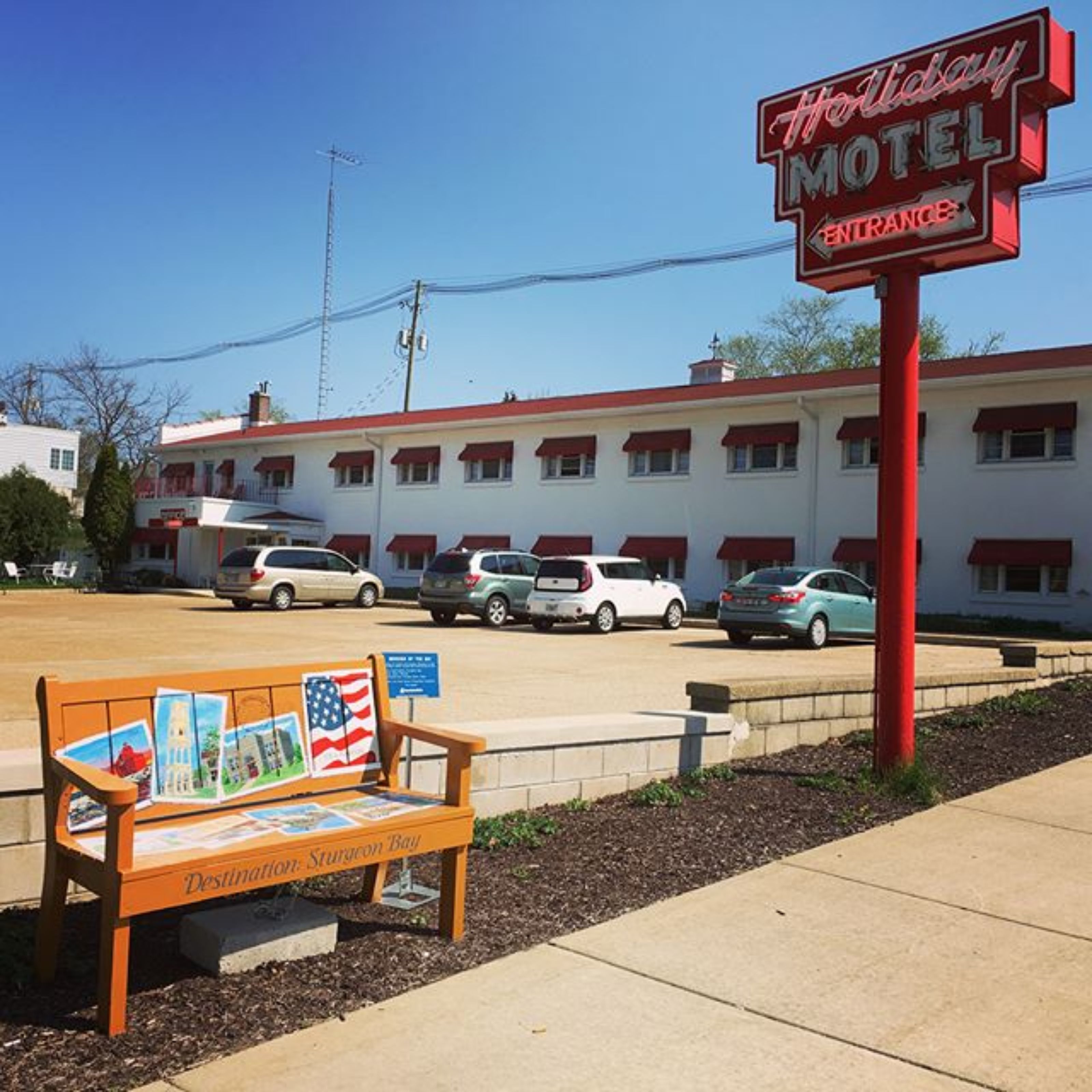 A Holiday Motel sign, a row of parked cars, and a bench with a map of Sturgeon Bay.