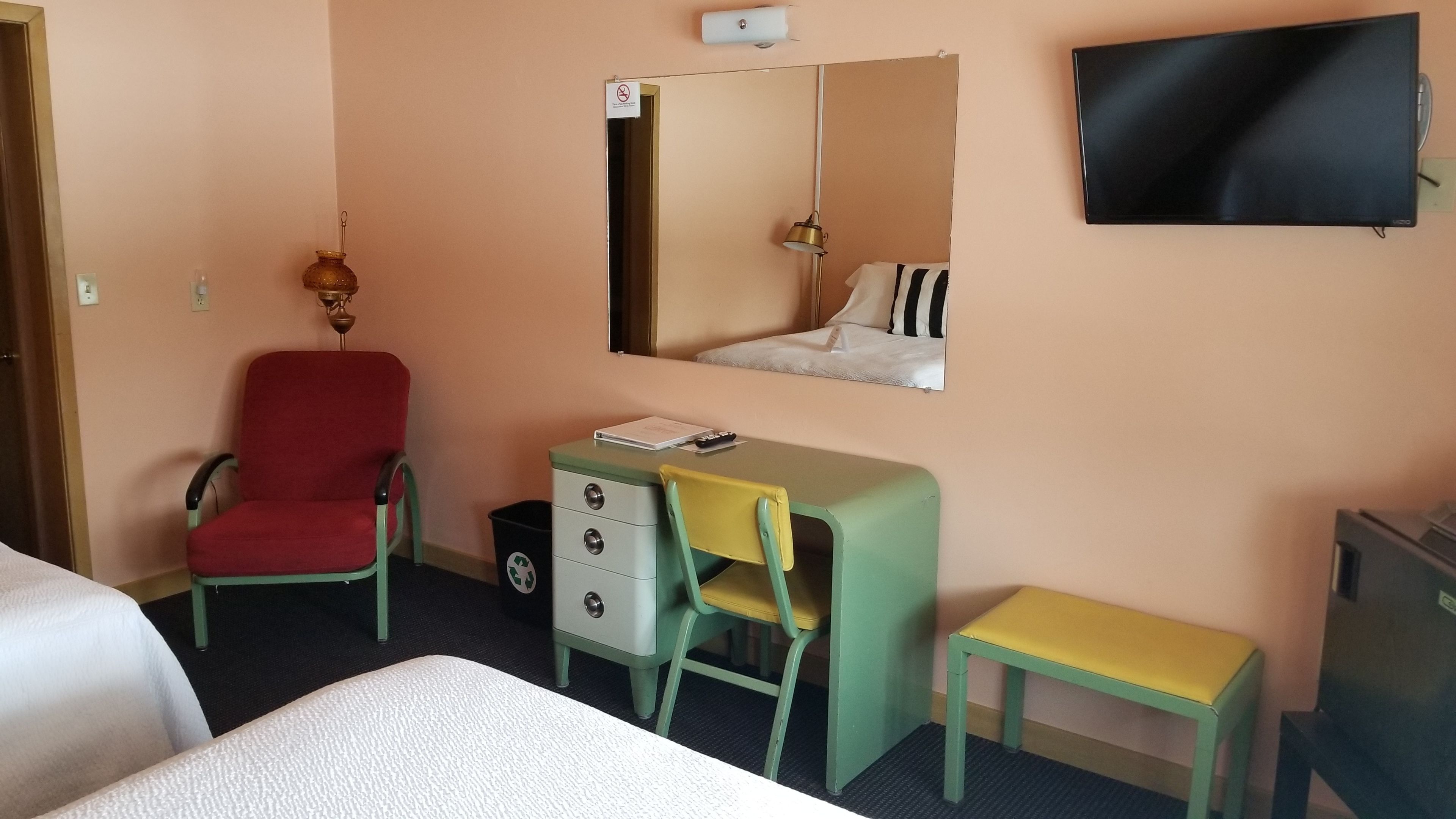 A hotel room with a bed, a desk with a chair, a small table, a trash bin, a television, and a mirror.