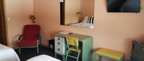 A hotel room with a bed, a desk with a chair, a small table, a trash bin, a television, and a mirror.