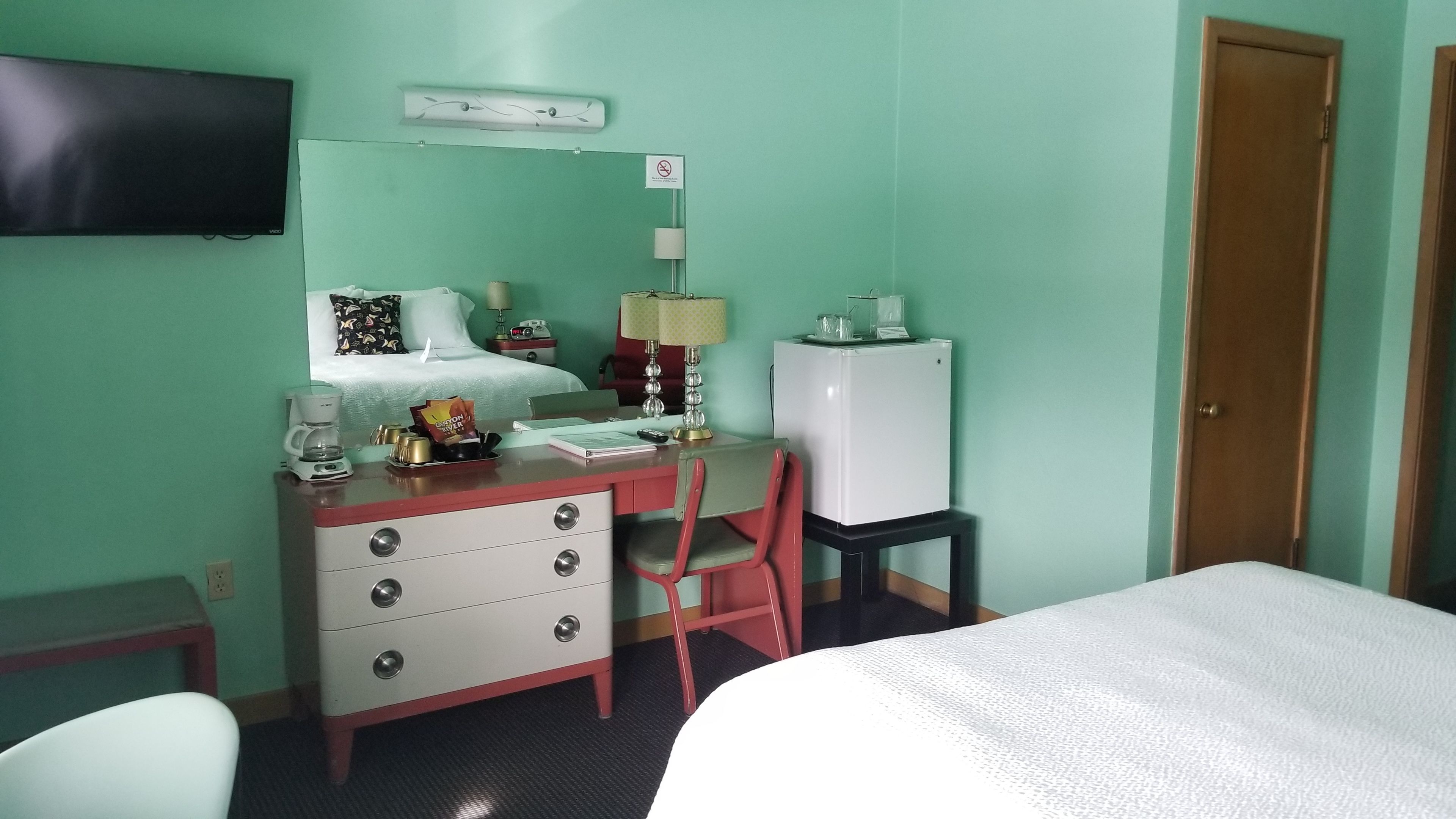 Habitación de hotel con cama, escritorio con silla, refrigerador pequeño, televisor montado en la pared y mesita de noche con lámpara.