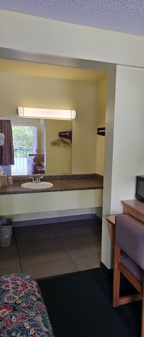 A hotel room with a bed, a bathroom with a sink and mirror, a wooden chair, and a television. - Travelers Inn (Gainesville)