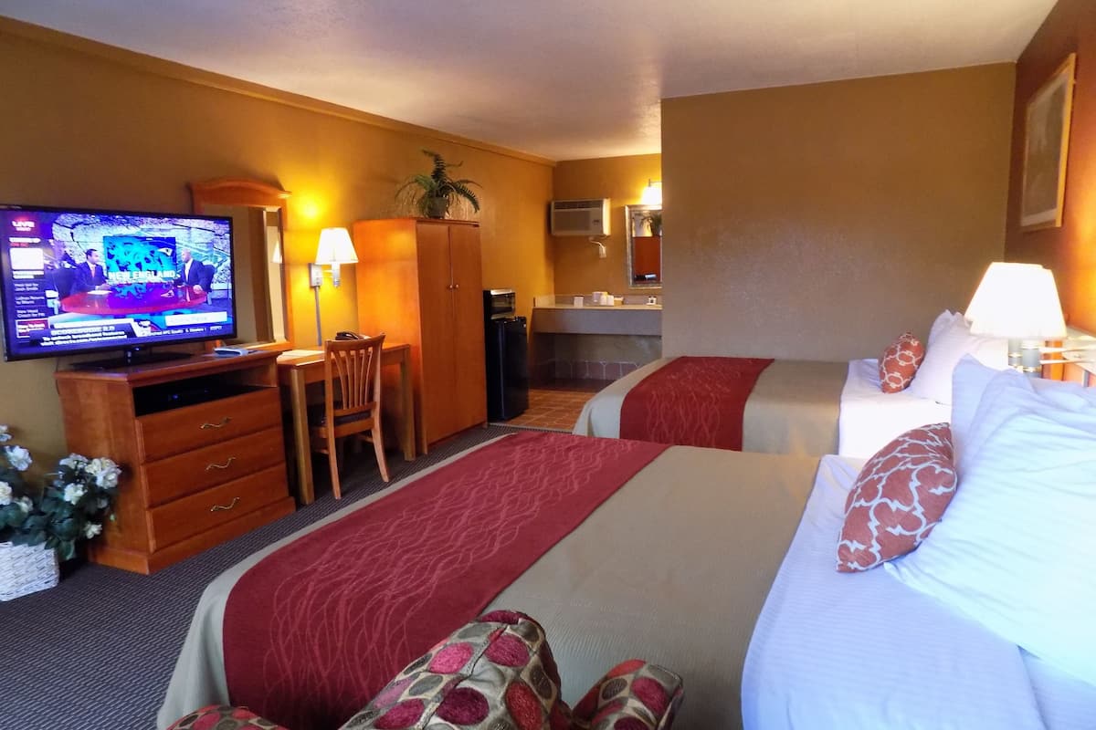 A hotel room with two beds, a television, a desk, a chair, a small kitchenette, and a bathroom.